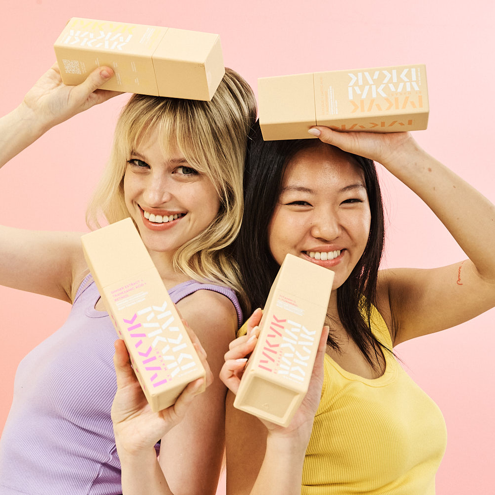 Two women holding makeup boxes on a pink background