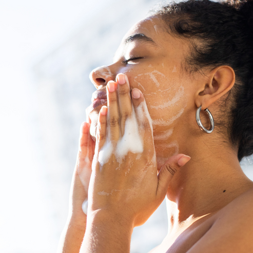 Woman applying Makari De Suisse Renewal Glow Cleansing Foam to her face with a blurred background